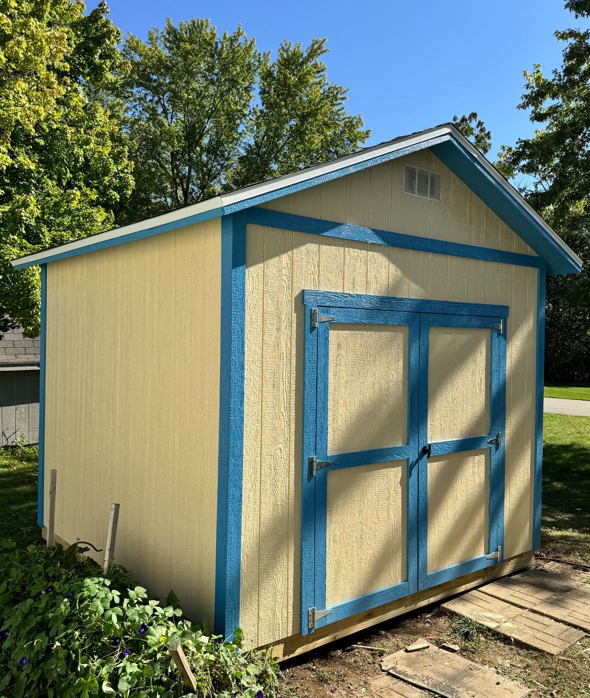 Tan and blue wooden shed in a garden with trees in the background under a clear blue sky.